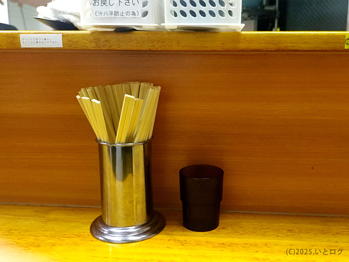 ラーメン二郎立川店の木製カウンターに割り箸と黒コップが整う。二郎 立川 ルールを守り静かに着丼を待つ準備シーン。