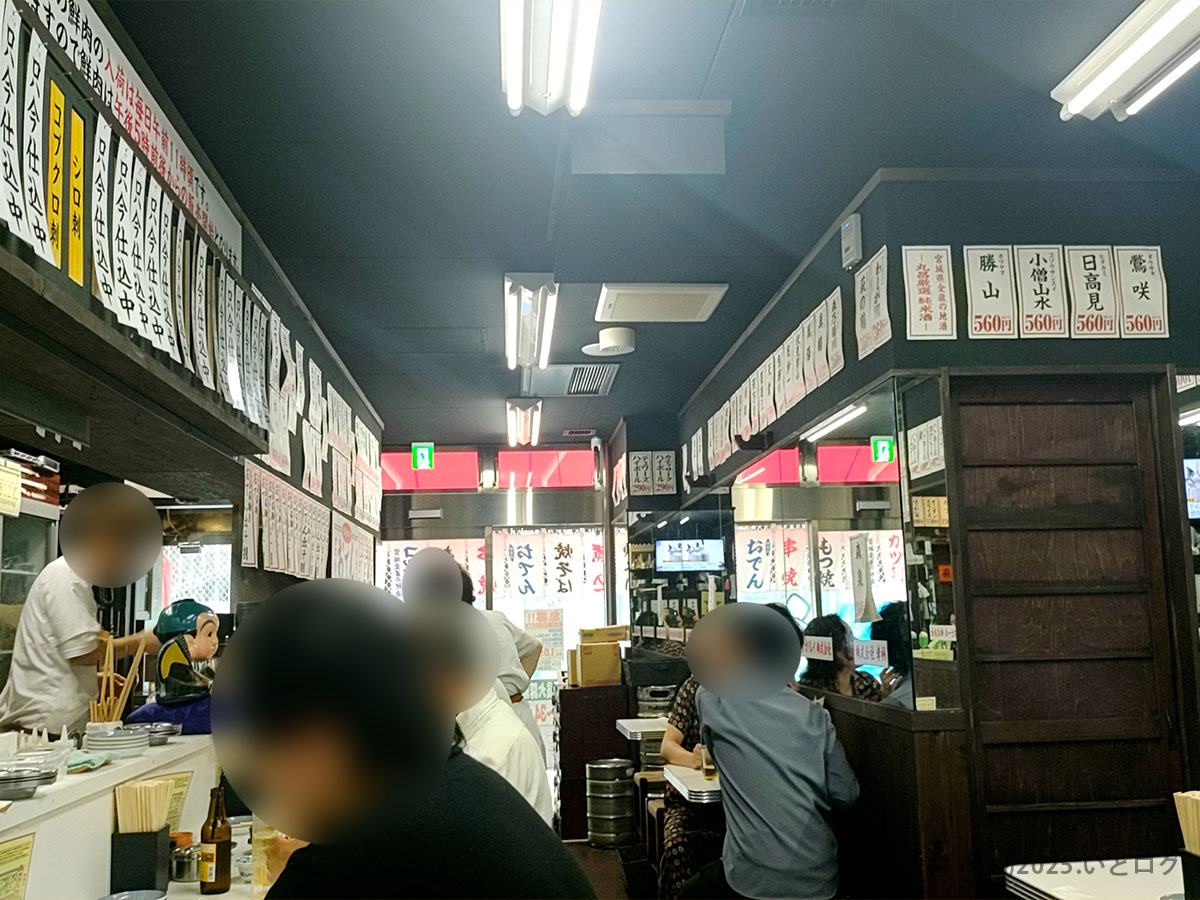 宮城県仙台市の大衆酒場『もつ焼き 丸昌 仙台』店内。壁一面に短冊メニューがずらりと並び、カウンターとテーブル席がお客さんで賑わう、昭和レトロな雰囲気