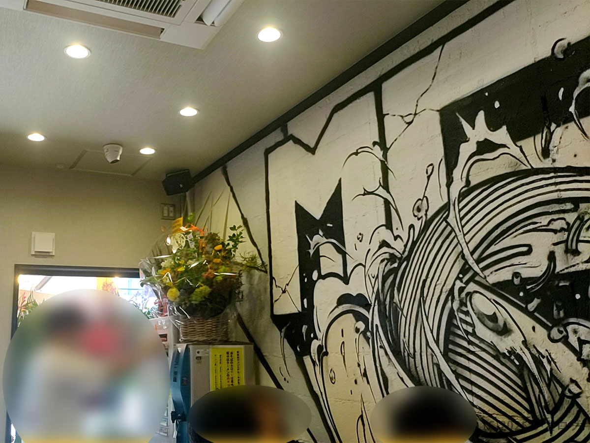 油そば専門店「東京麺珍亭本舗 甲府南店」の入口付近。麺を描いた迫力満点のモノクロ壁画と開店祝いの花が並び、竜王店に続く山梨2号店の雰囲気が伝わる。