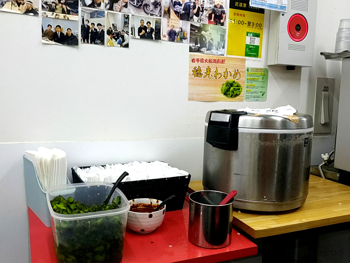 武道家 口(くにがまえ)国分寺のライス・漬物コーナー。名物のライス無料サービスやきゅうり漬けのセルフ提供が並ぶ。武道家系統ならではの“ライス必須の家系ラーメン文化”を表す一角。