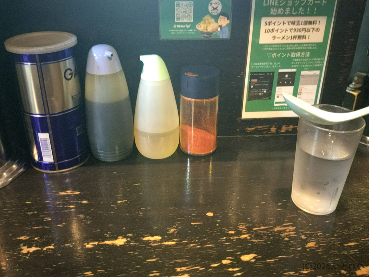 ラーメンマシンガン秋田店の卓上調味料とカウンター席の様子。二郎系らしいにんにくや唐辛子が並ぶ