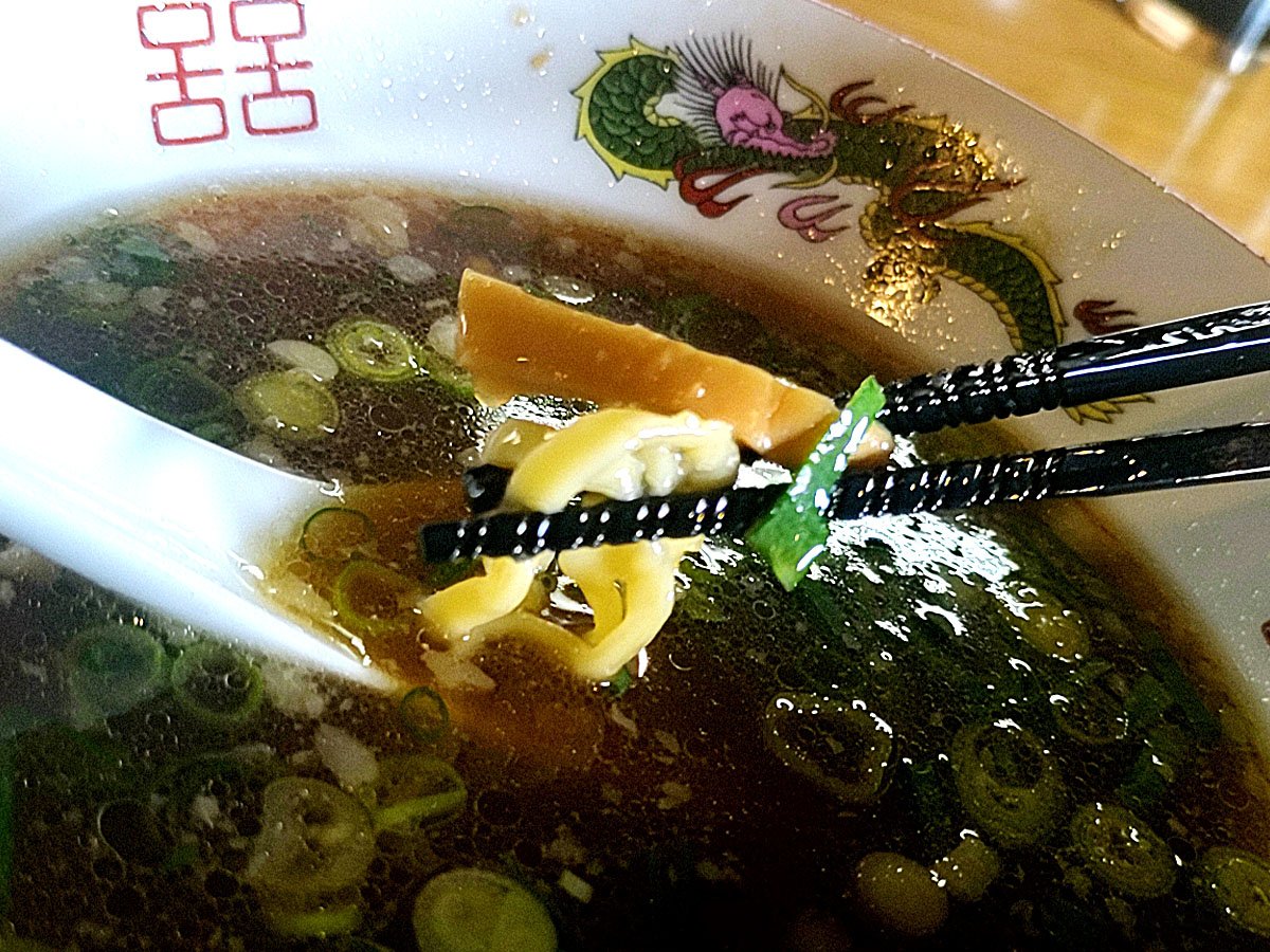 麺 㐂色（読み方は“きいろ”）のラーメン。黒い箸で持ち上げた縮れ麺とスープ、青ねぎの香りが食欲をそそる。
