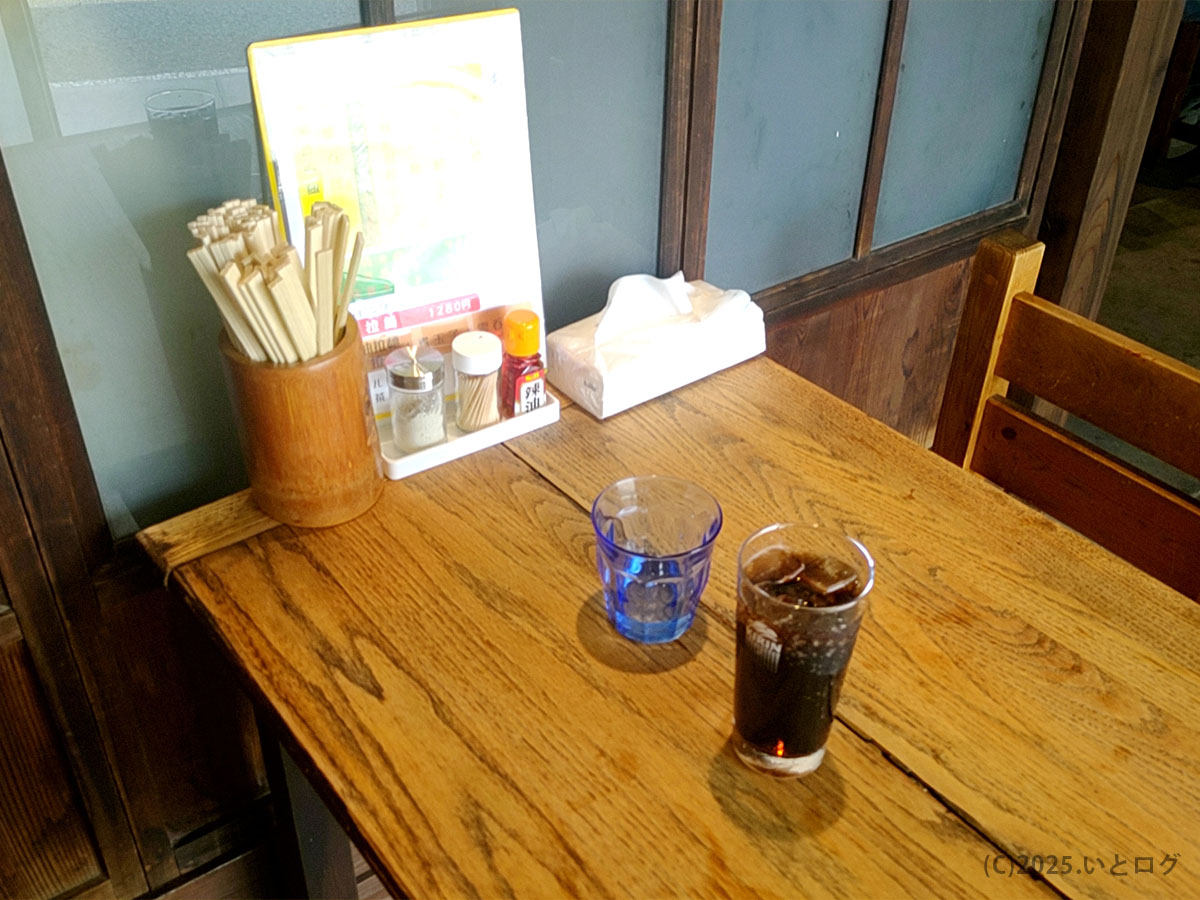 木の温もりある店内のテーブルに置かれたコーラと水。麺処 麗山（山梨県富士吉田市）の落ち着いた雰囲気を感じられる光景。富士吉田市ランチや観光途中の休憩にもぴったり。