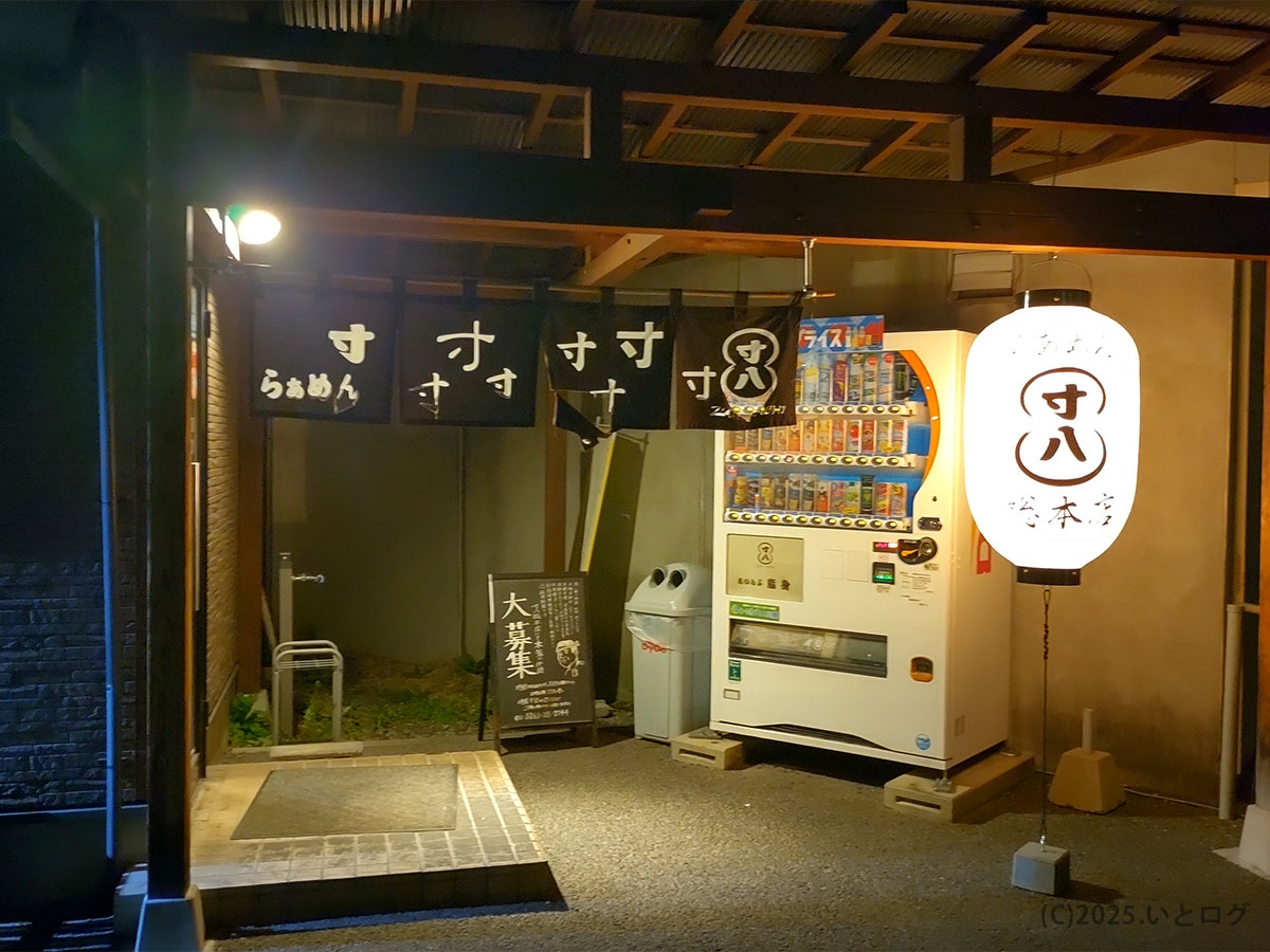 夜のらあめん寸八 総本店入口。提灯に「寸八」のロゴが灯り、店頭の寸八 自販機が松本の街角で客を迎えている情景。