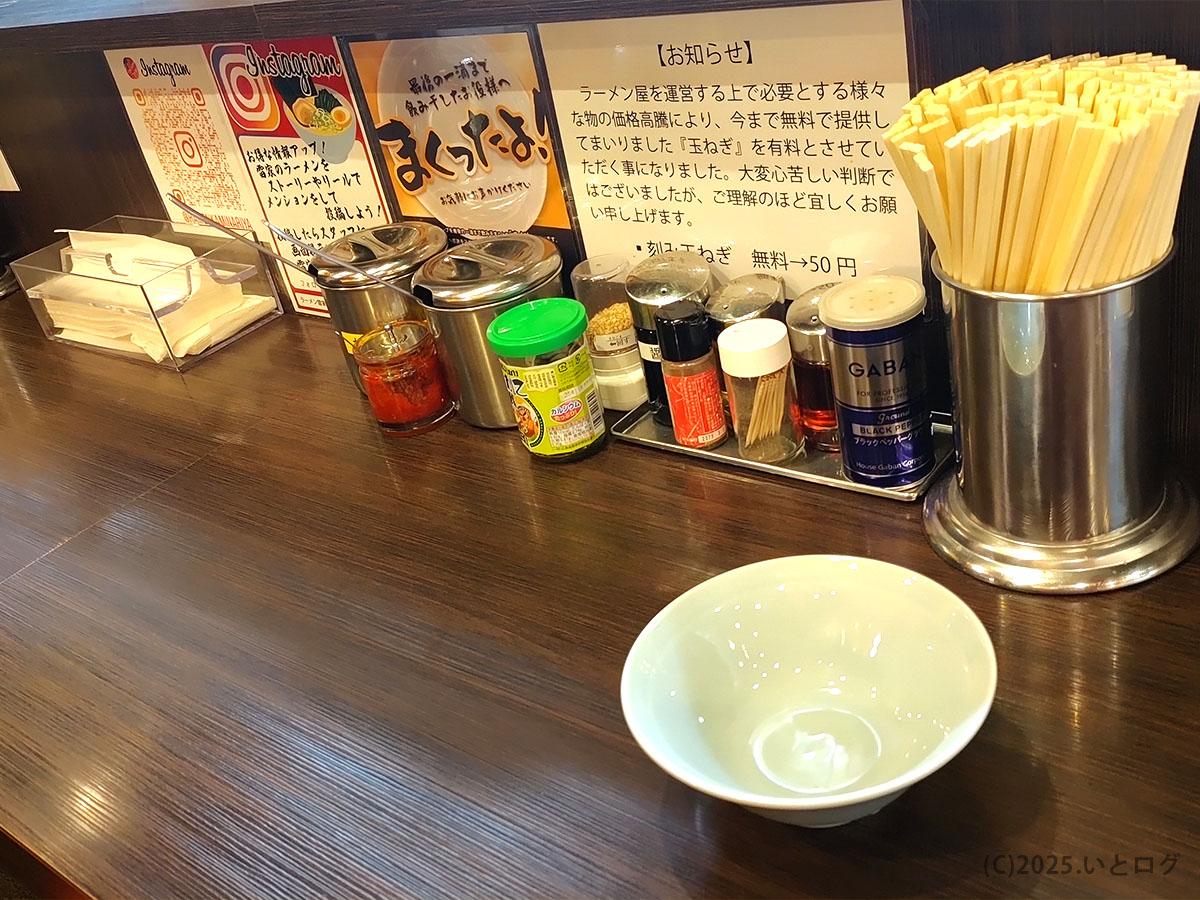 山梨・甲府市の「横浜家系 雷家 酒折」カウンター。無料紙エプロンや刻みニンニク、GABAN黒こしょうなど薬味がずらりと並び、雷家メニューのライス用ふりかけも見える。