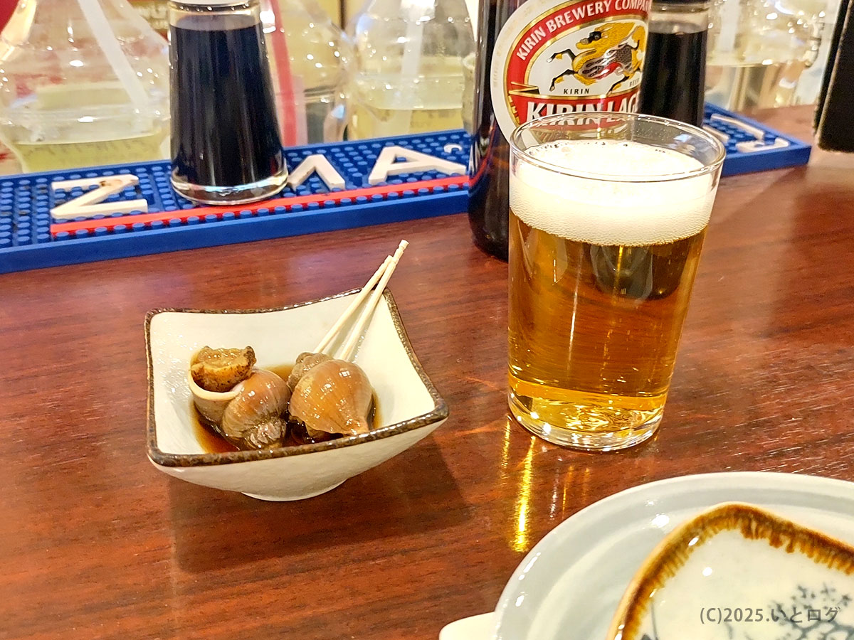 お通しのバイ貝煮とキリンラガー小グラス。まず一杯で“おきゃく”文化を体験するシーンを捉えたレビュー用カウンターショット。