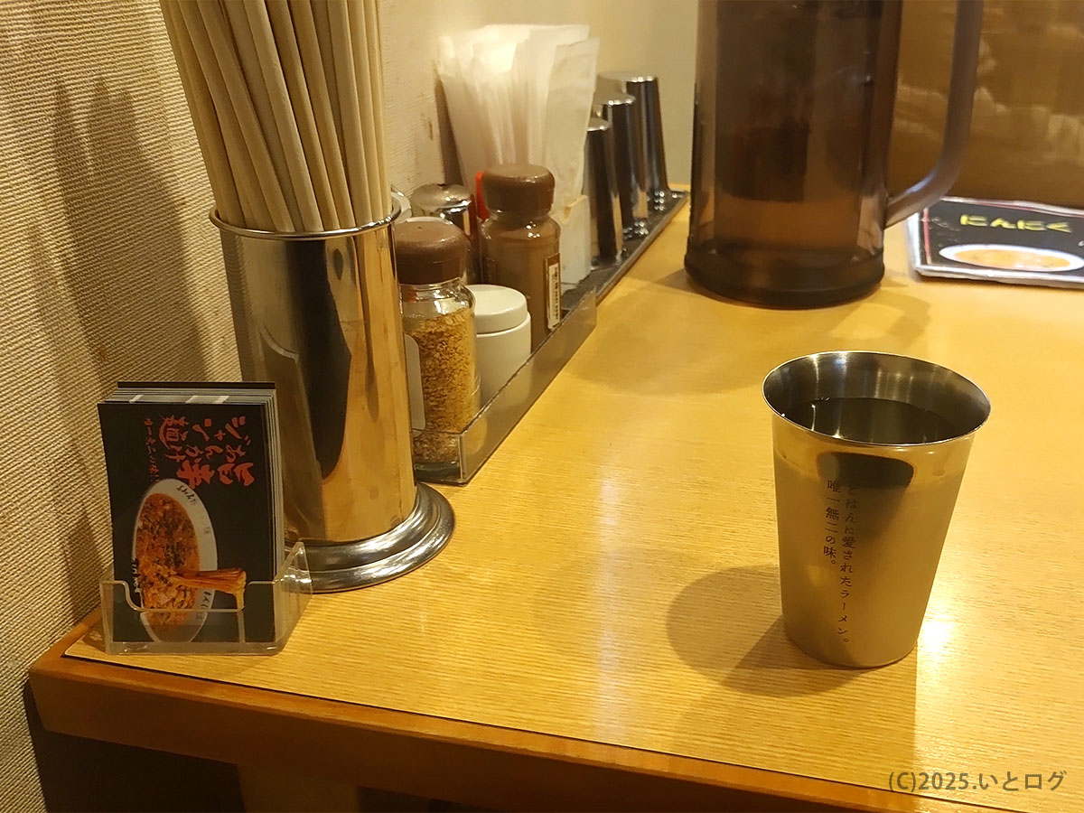 まんしゅう 高知 本店カウンター。箸立てと調味料、ステンレスカップに刻まれた「ごはんに愛されたラーメン」のフレーズが映える