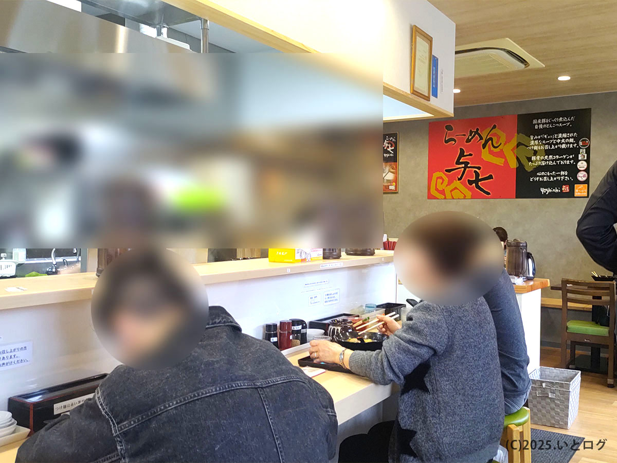 滋賀県大津市にあるらーめん与七 堅田店の店内風景。カウンター席で豚そばを食べるお客さんの背後には赤と黒の大きな店名看板が掲げられ、駐車場が広く事故の心配なく利用できるのも魅力。