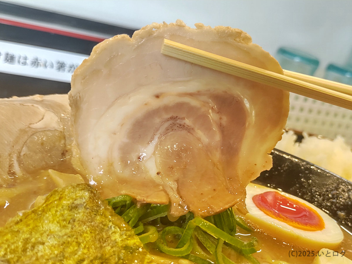 ラーメン与七の豚そばに乗る大きなバラロールチャーシューを箸で持ち上げたところ。脂身と赤身の渦が美しく、下には海苔や青ねぎ、半熟玉子が見える。