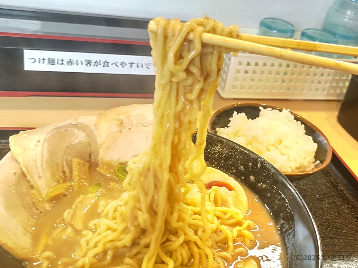 もちもちした中太縮れ麺を箸で持ち上げた瞬間。濃厚スープがよく絡んだラーメン与七 豚そばはご飯との相性も良く、後ろにはライスの小鉢も写っている。