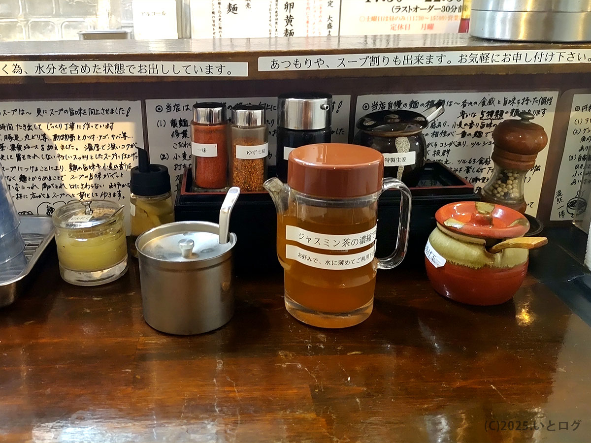つけ麺紅葉 国分寺店のカウンター調味料。ゆず七味や特製にんにく、ジャスミン茶の濃縮液など個性的なラインナップが並ぶ。ラーメンもみじらしい自由な味変スタイル。