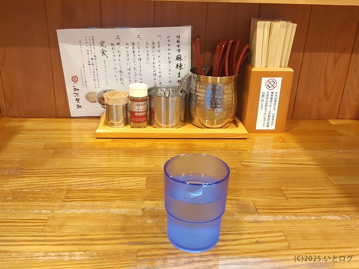 甲府ラーメン『初志貫徹』2号店の木目カウンターに水コップと調味料が整列し、ツイッターでも映える落ち着いた店内を写す。