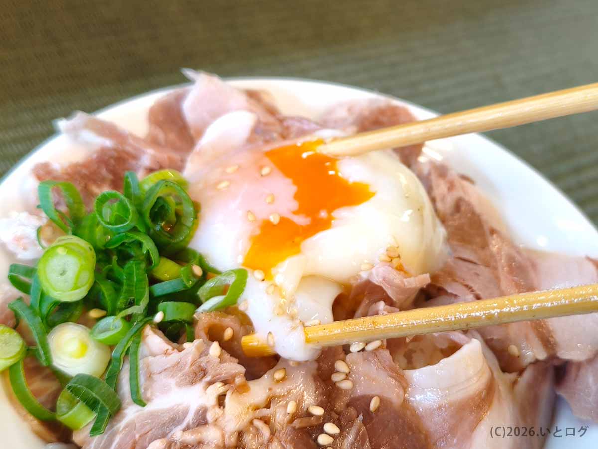 伊吹いりこセンターのチャーシュー丼にのった温玉を箸で割る様子。刻みネギや白ごま、たっぷりのチャーシューが盛られている