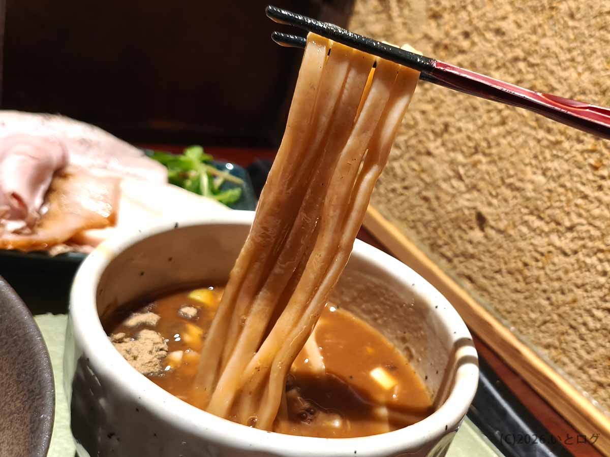 つけ麺 繁田の幅広い自家製麺を、魚介の風味が漂う濃厚なつけ汁につける様子