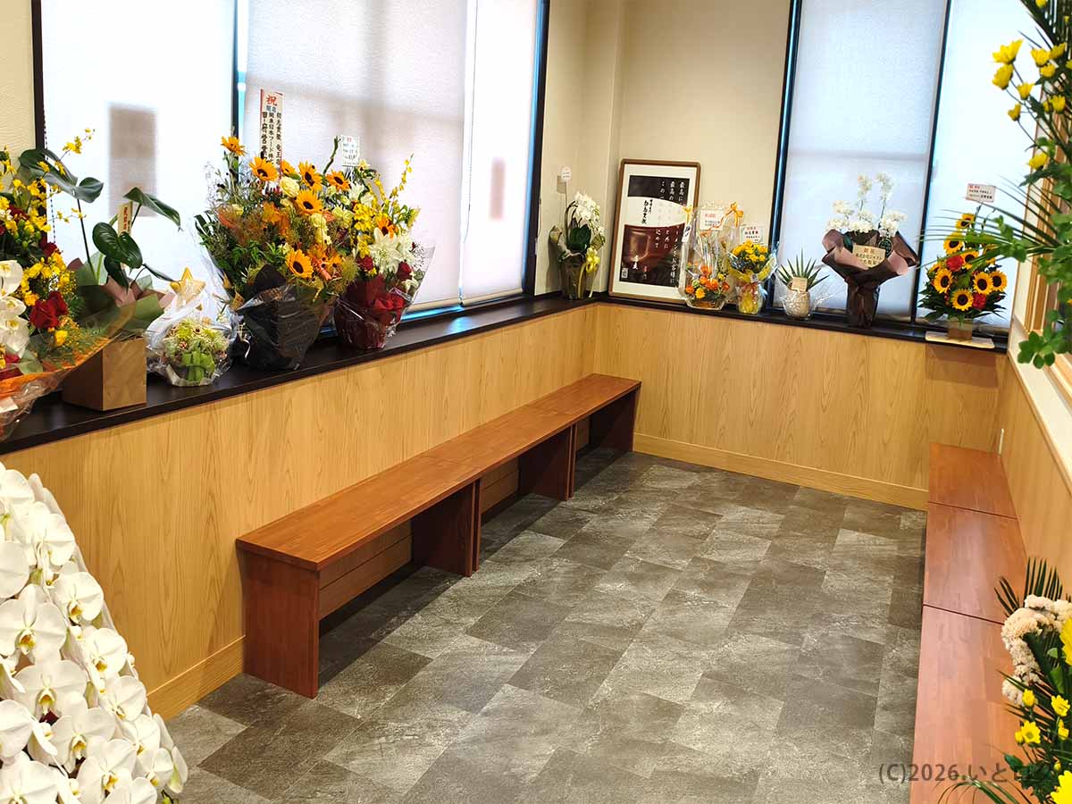 初志貫徹 甲斐竜王店の店内に設けられた、開店祝いの花に囲まれた木製ベンチ付きの待合スペース