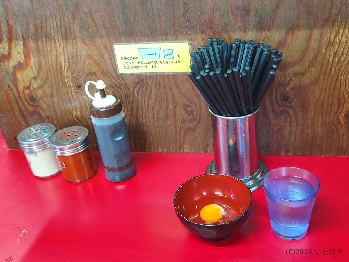 ラーメン二郎 新潟店の赤いカウンター。卓上調味料や箸、水、生たまごを入れた器が並ぶ店内の様子。