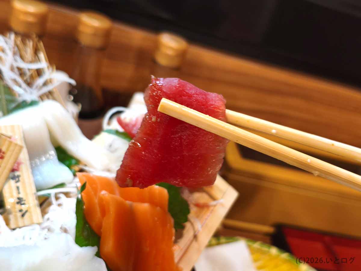 帯広の魚の一心で注文した刺身8品盛りの本マグロ。箸で持ち上げた赤身の切り身と、奥に盛り付けられたサーモンなどの刺身。
