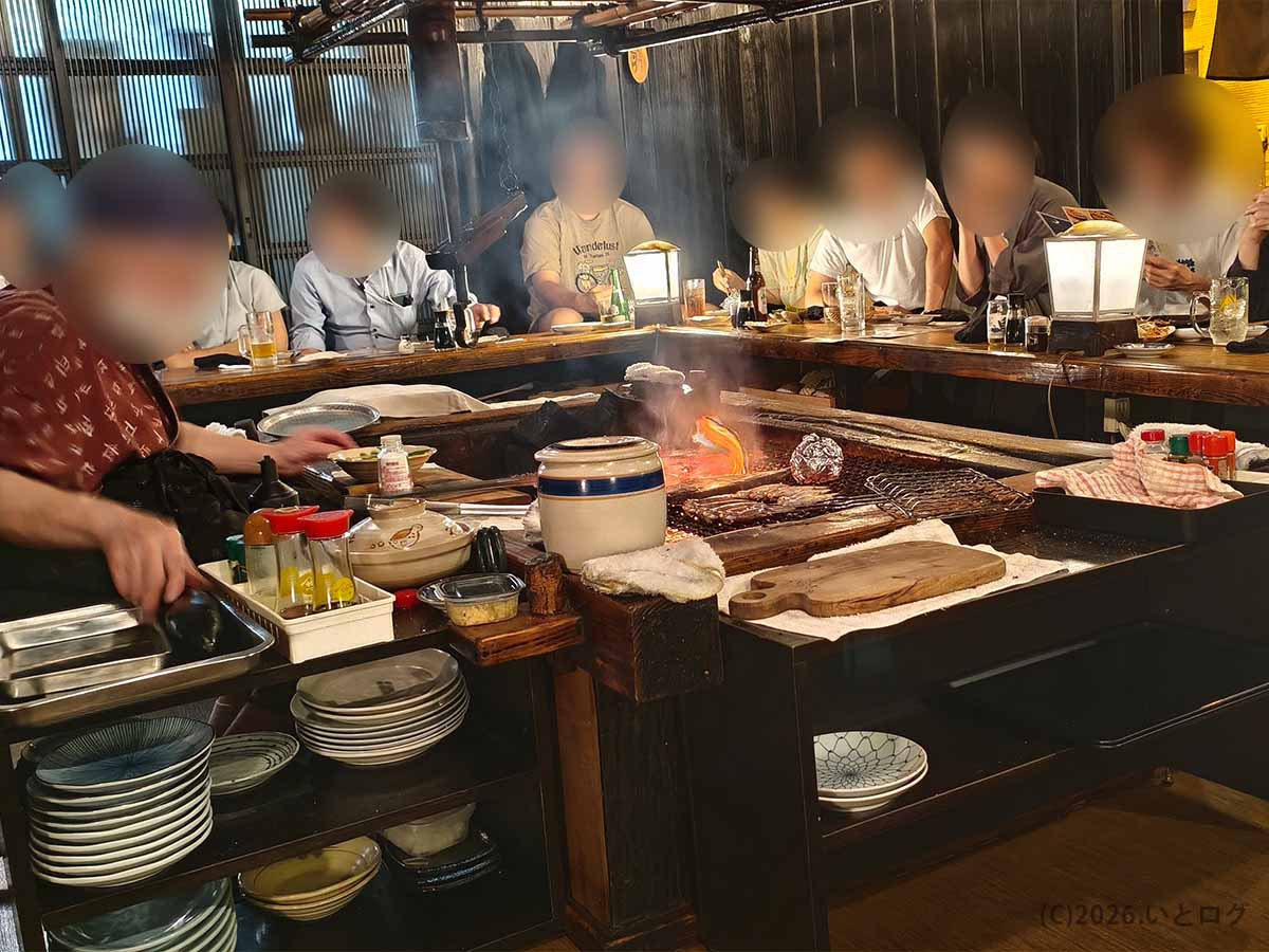 炉端焼き発祥の店「炉ばた」の中央にある炭火の囲炉裏。焼き網の上で食材が焼かれ、周囲のカウンター席で客が食事を楽しんでいる