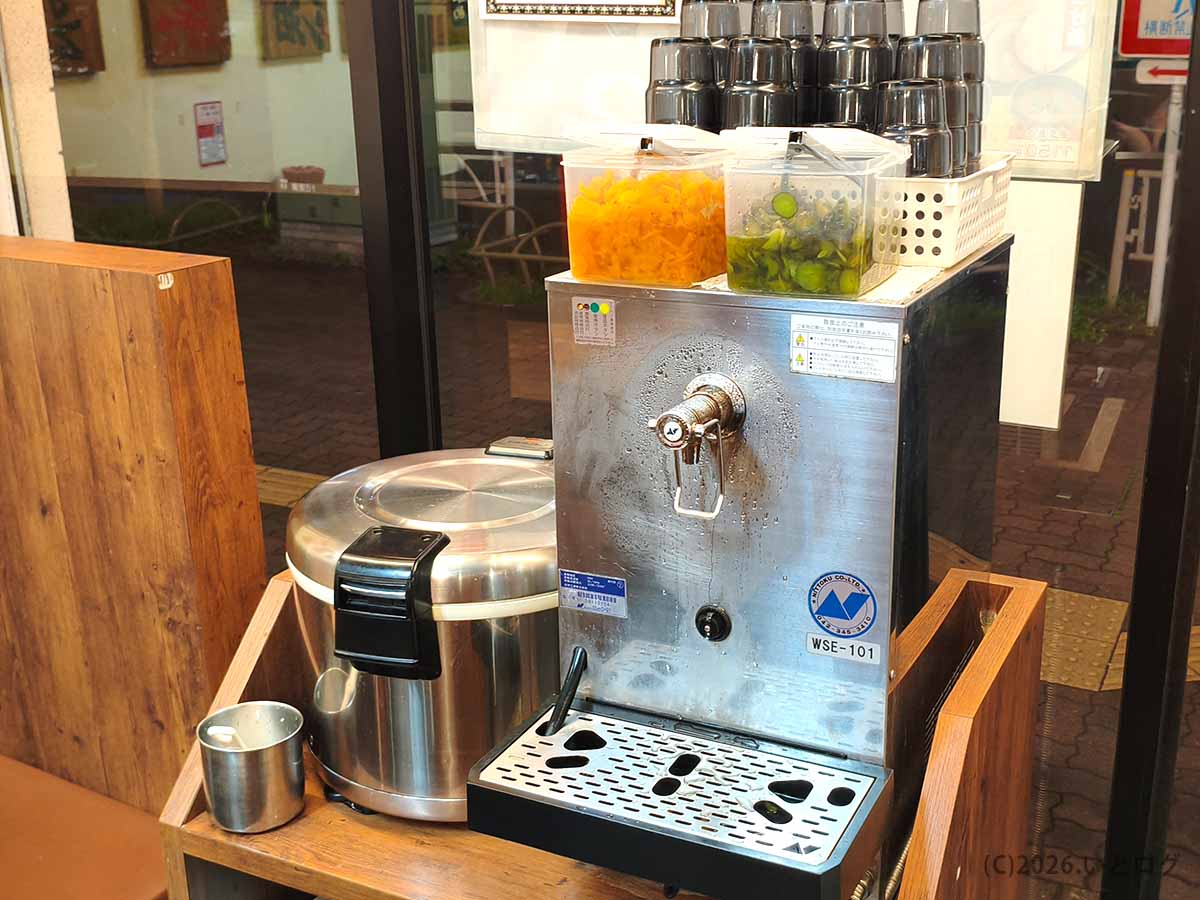 巣鴨家の終日無料ライス食べ放題コーナー。ご飯の保温ジャー、給水機、漬物が置かれたセルフサービススペース