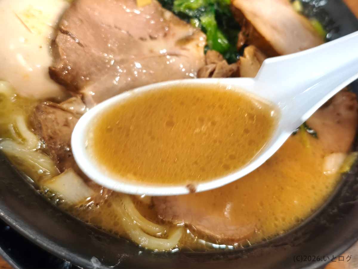 巣鴨家の家系ラーメンのスープを白いレンゲですくった様子。茶褐色の豚骨スープとチャーシューが写る