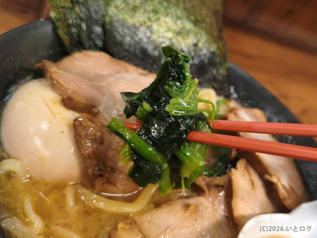 巣鴨家の豚骨ラーメンにのったほうれん草を箸で持ち上げた様子。奥にはチャーシュー、味玉、海苔が見える
