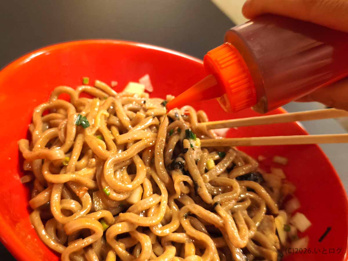 甲府の油そば専門店あぶらギルドで、タレの絡んだ太麺に卓上のラー油を加える様子。酢に続いて辛味を調整しながら味変を楽しむ。