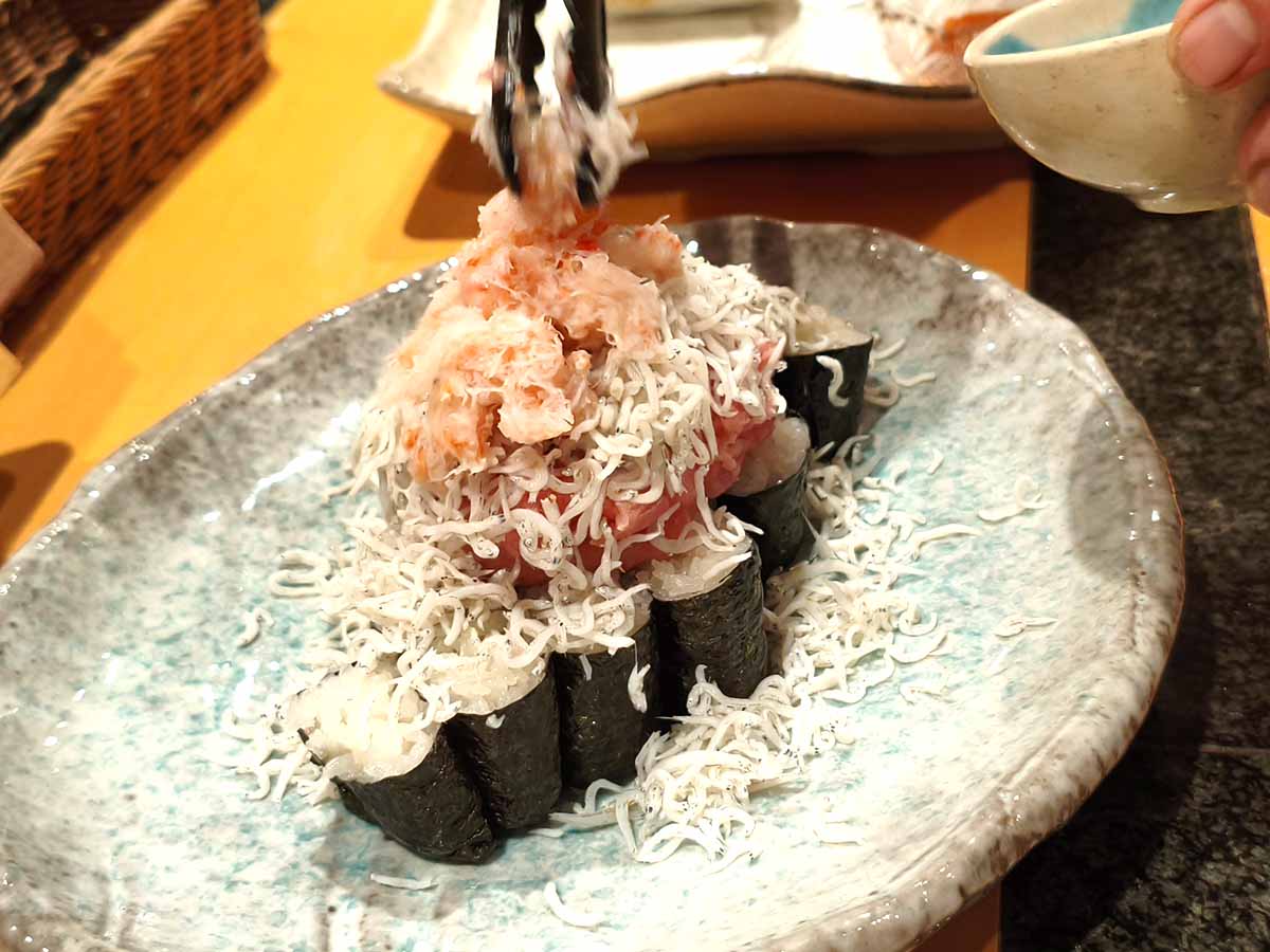 海産物こぼれ巻きの盛り付け途中。海苔巻きの上にネギトロ、しらす、ほぐしたカニの身が重なり、トングで具材を追加している