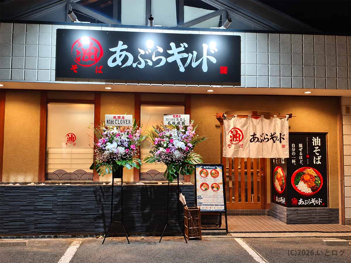 2026年10月21日に山梨県甲府市朝気にオープンした油そば専門店「あぶらギルド」の夜の外観。黒い看板と白い暖簾が目印で、開店祝いの花が並んでいる。