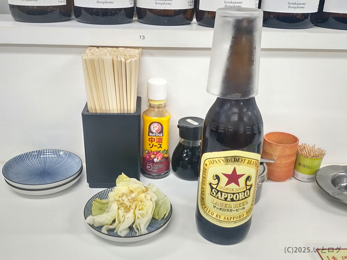 サッポロ赤星ラガービールと卓上キャベツが並ぶ『もつ焼き 丸昌 仙台』のカウンター。ソースや醤油など調味料もそろい、ひとり飲みでも落ち着ける酒場の風景