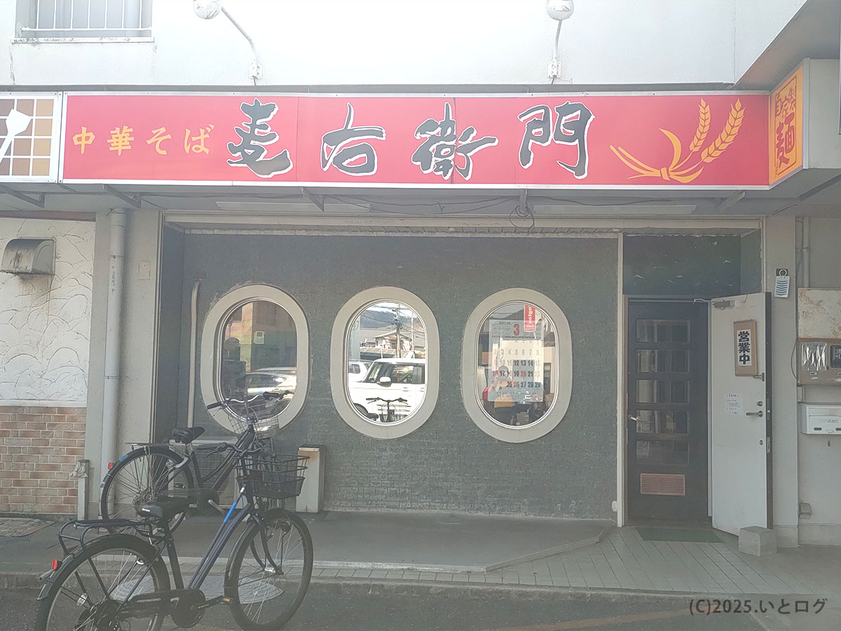 赤い看板に「中華そば 麦右衛門」と筆書きが映えるレトロ外観。楕円窓の奥に客の姿がちらり。店舗前には数台分の共用スペースがあり、中華そば麦右衛門 駐車場の目安になる。