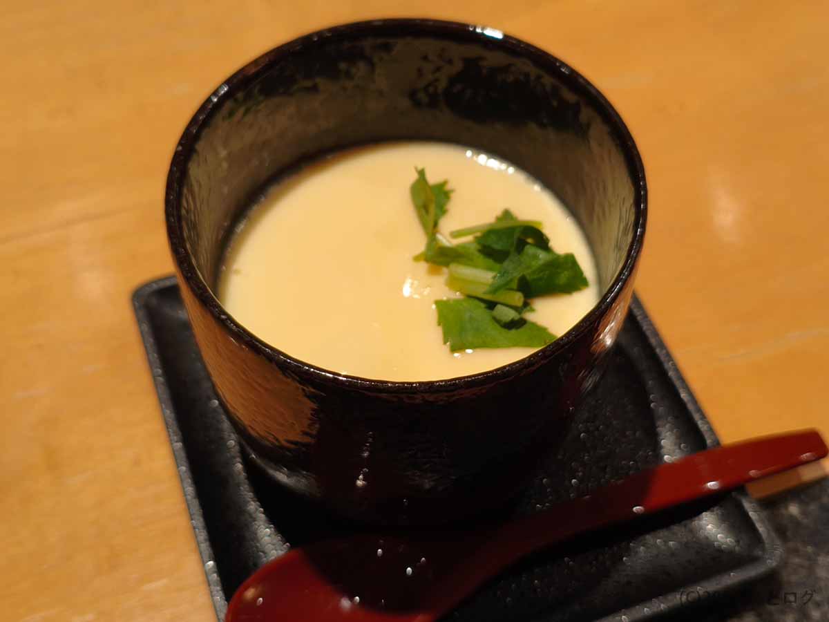 二代目たわらの茶碗蒸し。黒い陶器の器に入った淡い黄色の卵生地の上に三つ葉が添えられ、黒い角皿と赤いスプーンがセットされている
