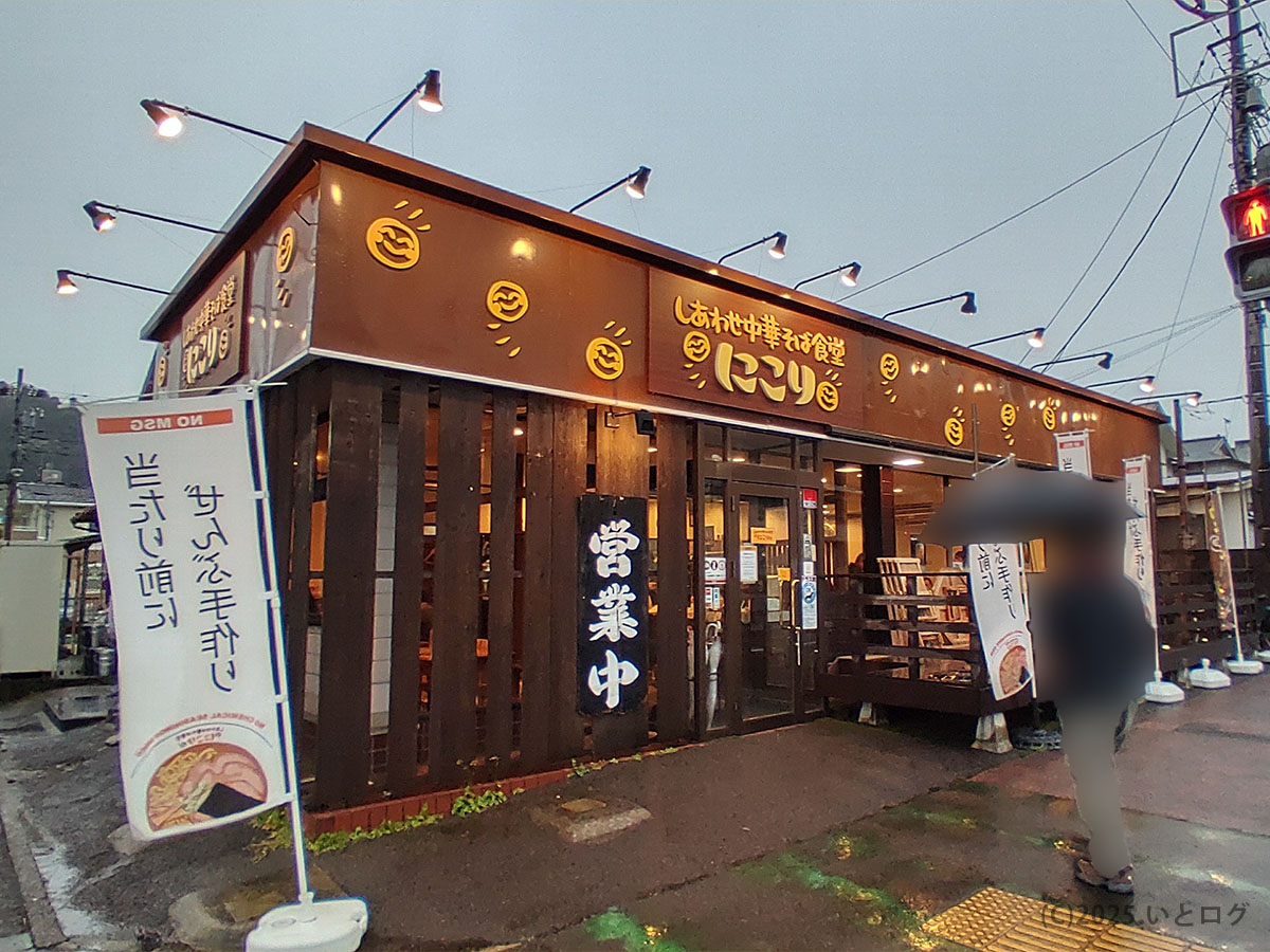 湯河原・しあわせ中華そば食堂にこりの外観。にっこりマークの茶色い木造店舗がライトに照らされ、のぼり旗には手打ち麺推しの文言。雨上がりでも「営業中」の文字が誘う。