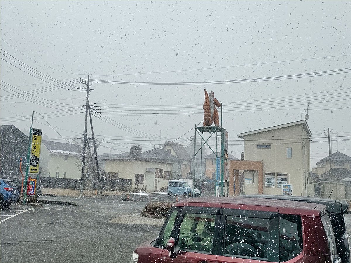 雪が舞う手打 焔の駐車場とラーメン焔←案内看板。鯉のオブジェが目印で、待ち時間・並び状況も分かる冬景色ショット