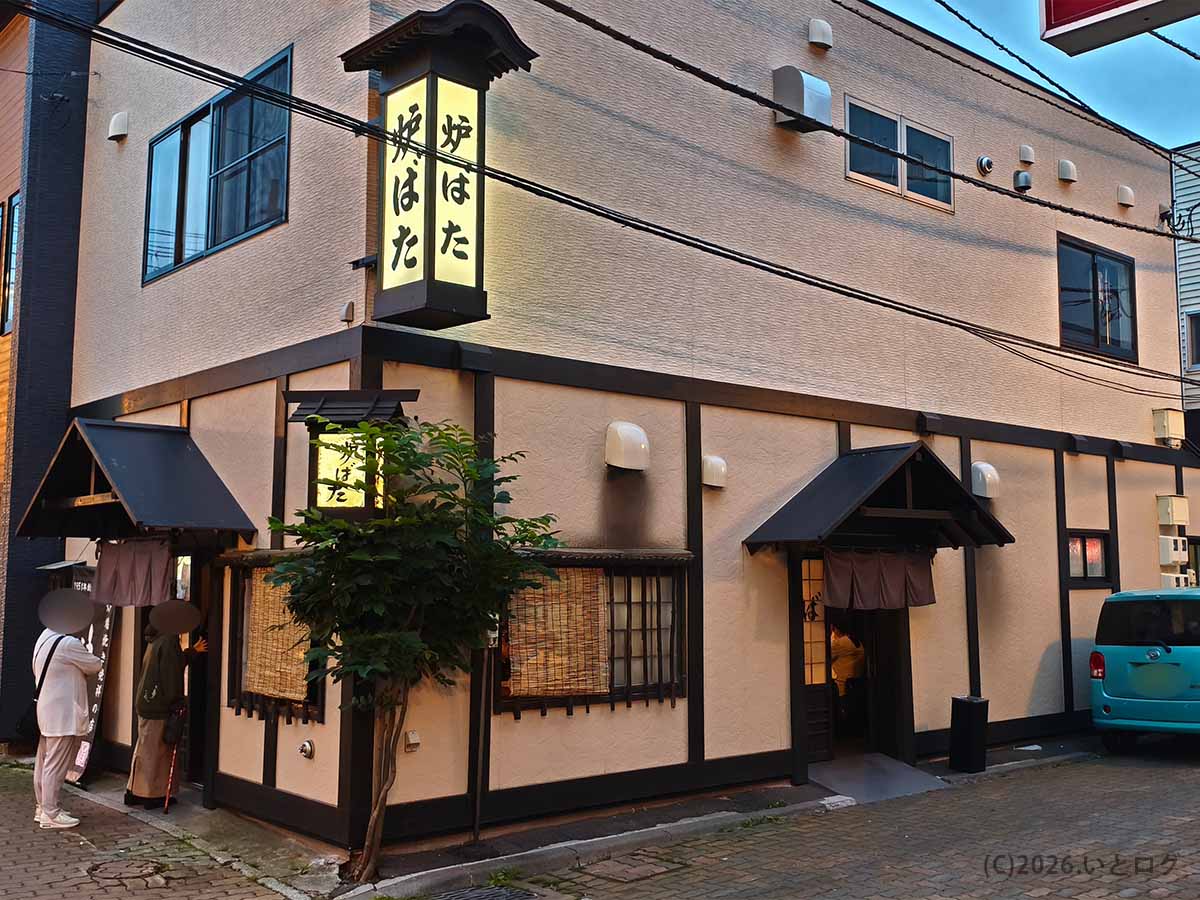 北海道釧路市栄町にある炉端焼き店「炉ばた」の外観。白い外壁の建物に店名が書かれた縦長の看板と暖簾が設置されている