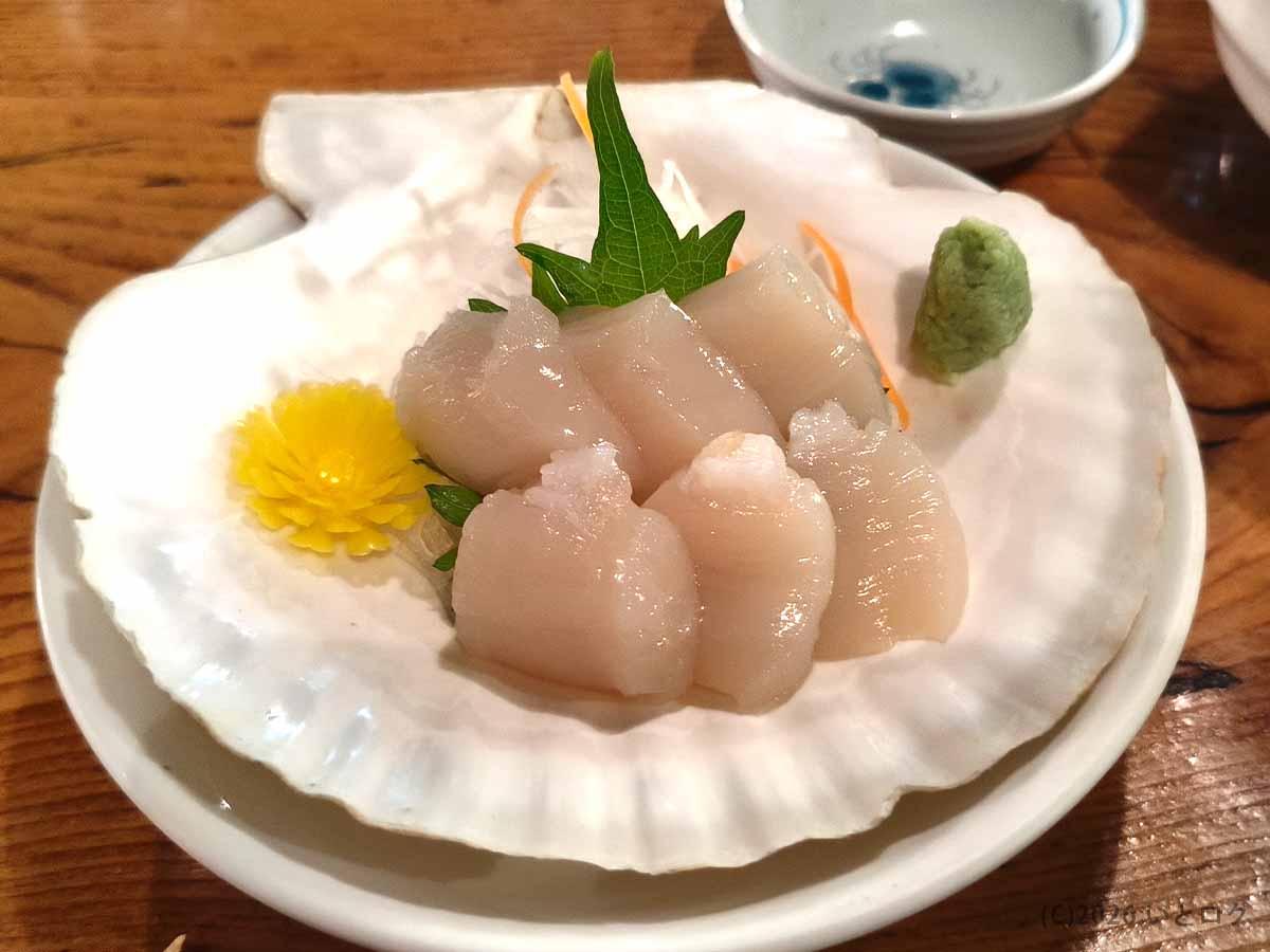 炉ばたで提供されたホタテの刺身。大きな貝殻を器にして、厚切りのホタテ貝柱が大葉やワサビとともに盛り付けられている